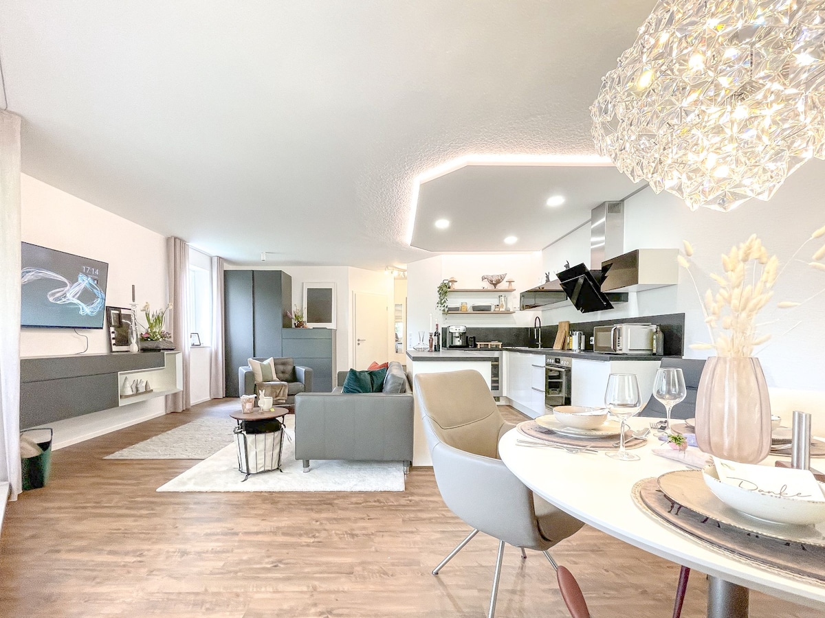 An open living space includes a cozy seating area with a gray sofa and a round dining table set for four. A modern kitchen is visible in the background, featuring sleek cabinetry and updated appliances. Natural light brightens the room, enhancing the contemporary décor.