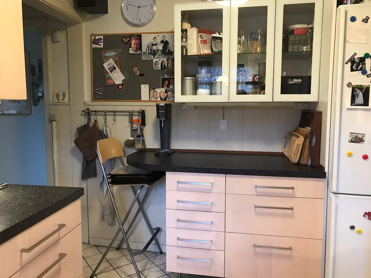 A functional kitchen corner features a compact folding chair positioned beside a black countertop. The countertop is accompanied by cabinets showcasing glassware and organized kitchen items. A bulletin board displays photographs and notes, contributing to a homely atmosphere.