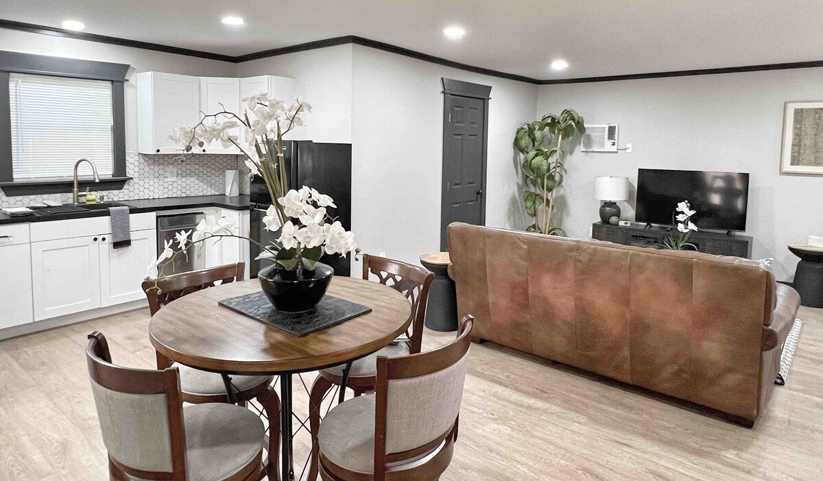 A contemporary living space is highlighted by a round dining table with plush chairs, accented by a decorative vase filled with white flowers. The open kitchen features sleek cabinetry and modern appliances, while a comfortable sofa and a smart TV create an inviting lounge area.