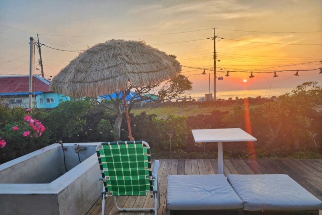 A wooden deck area is presented, featuring a sunshade and lounge chairs. A small table stands nearby, complementing the cozy outdoor setting. In the background, a vibrant sunset illuminates the sky, reflecting on the tranquil waters, while blooming roses add a touch of nature.