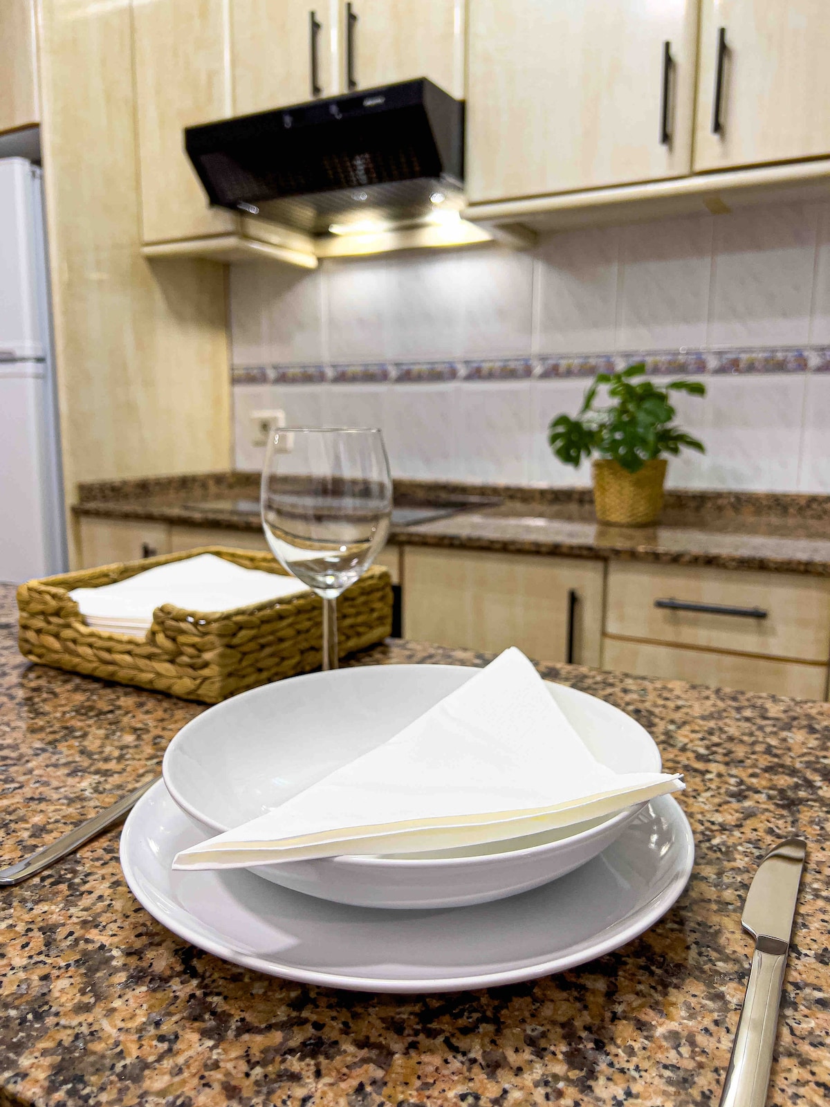A kitchen countertop features granite surfaces with neatly arranged plates, utensils, and a glass. A small potted plant adds a touch of greenery. A woven basket holds napkins, and an overhead range hood is seen in the background.