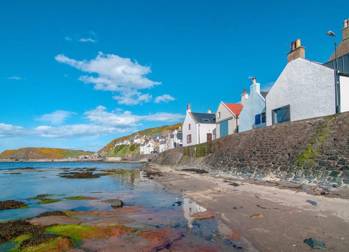 The coastline features a view of charming white cottages lining the shore, set against a backdrop of lush green hills and a clear blue sky. The calm waters reveal colorful seaweed and rocky outcrops along the sandy beach.