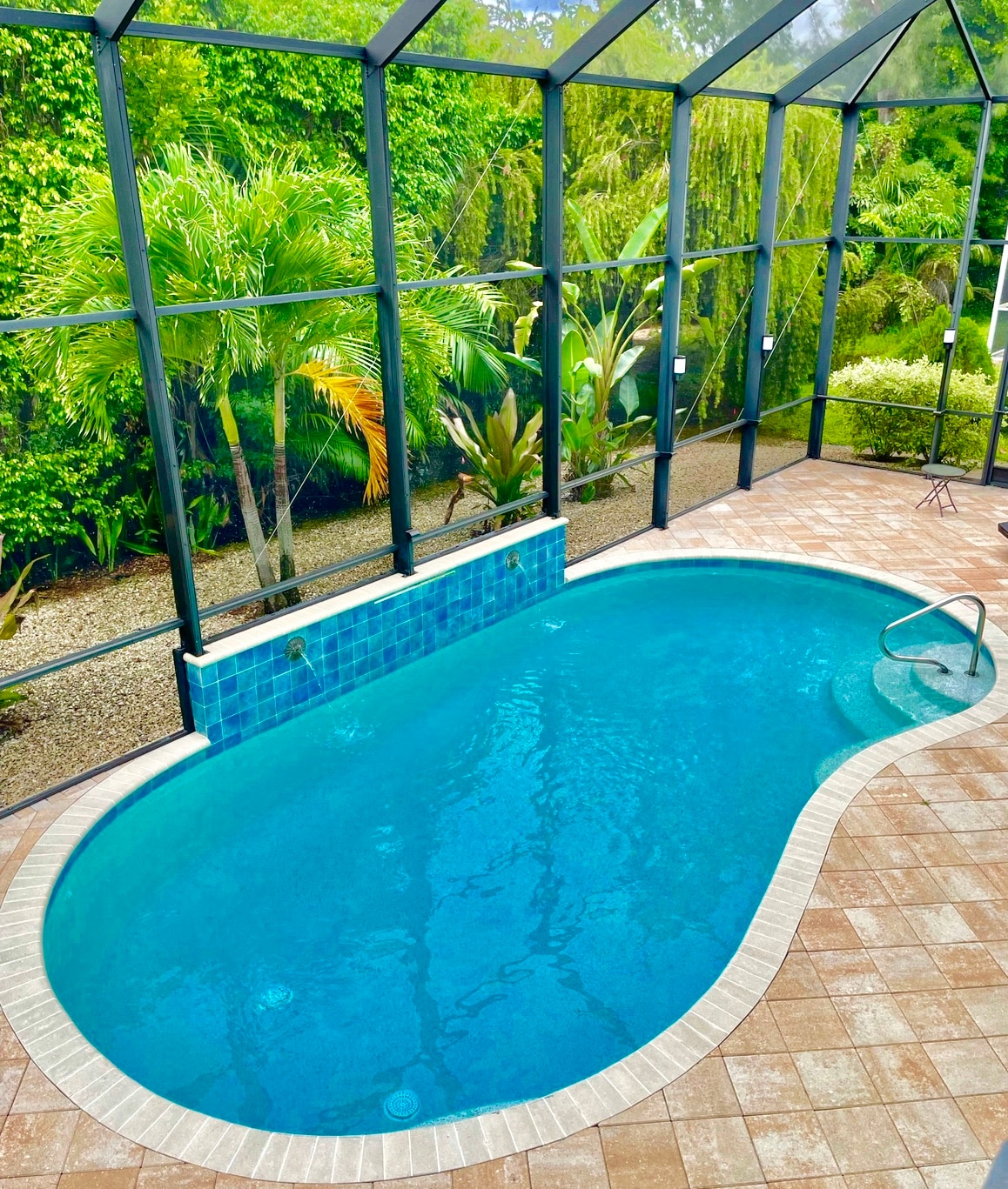 A private pool is enclosed by a glass screen, allowing views of the surrounding tropical plants. The pool features a tiled back wall and a curved edge, set on a patio with textured pavers. Lush greenery is visible beyond the enclosure.