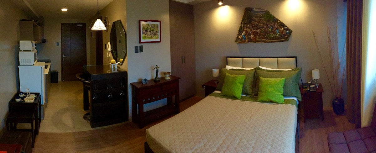 The studio unit features a double-sized bed adorned with soft green accent pillows. A modest kitchen area is visible in the background, alongside a wooden dining table with bar chairs. Artistic wall decor adds character, complemented by warm lighting and a spacious layout.