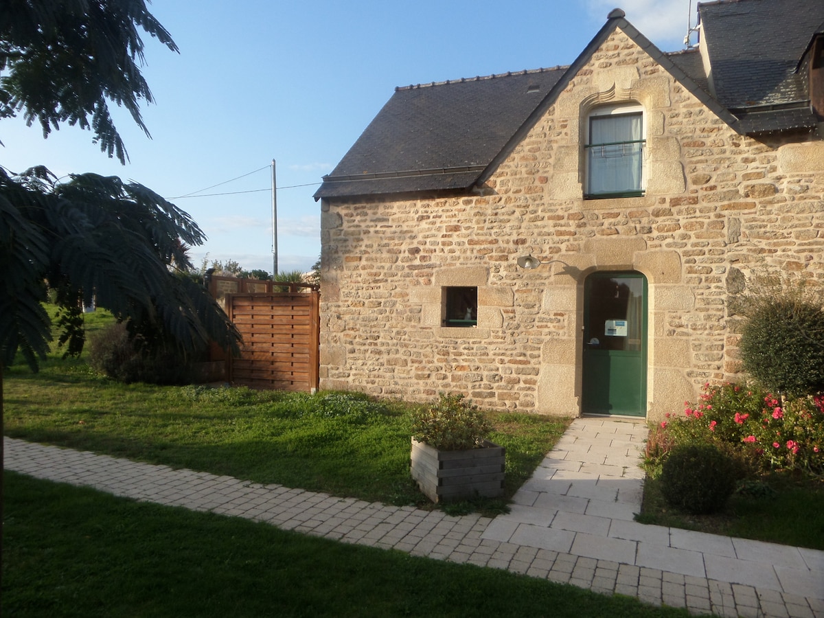 The exterior of the stone cottage is presented with a green door and a window beneath a gentle slope of the roof. A pathway lined with blooming flowers leads to the entrance, while a grassy area and a wooden fence are visible in the background.