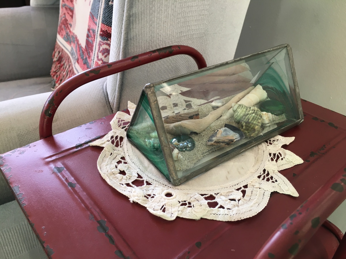 A glass display case containing assorted seashells and beach artifacts is placed atop a delicate lace doily on a red metal side table. A cozy armchair with a patterned throw can be seen in the background, enhancing the inviting feel of the space.