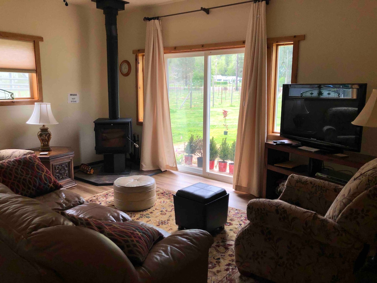 A comfortable living room features a cozy sofa and armchairs across from a wooden television stand. Large sliding glass doors provide a view of the outdoor greenery. A wood-burning stove adds warmth to the space. Soft curtains frame the windows, enhancing the natural light.