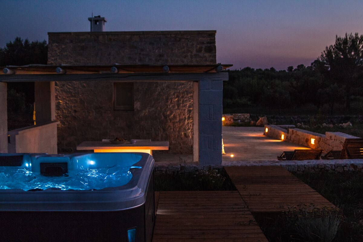 A hot tub filled with illuminated water is positioned on a wooden deck, offering a relaxing ambiance. The surrounding area features a stone structure and outdoor seating, with gentle lighting enhancing the tranquil atmosphere as dusk sets in.