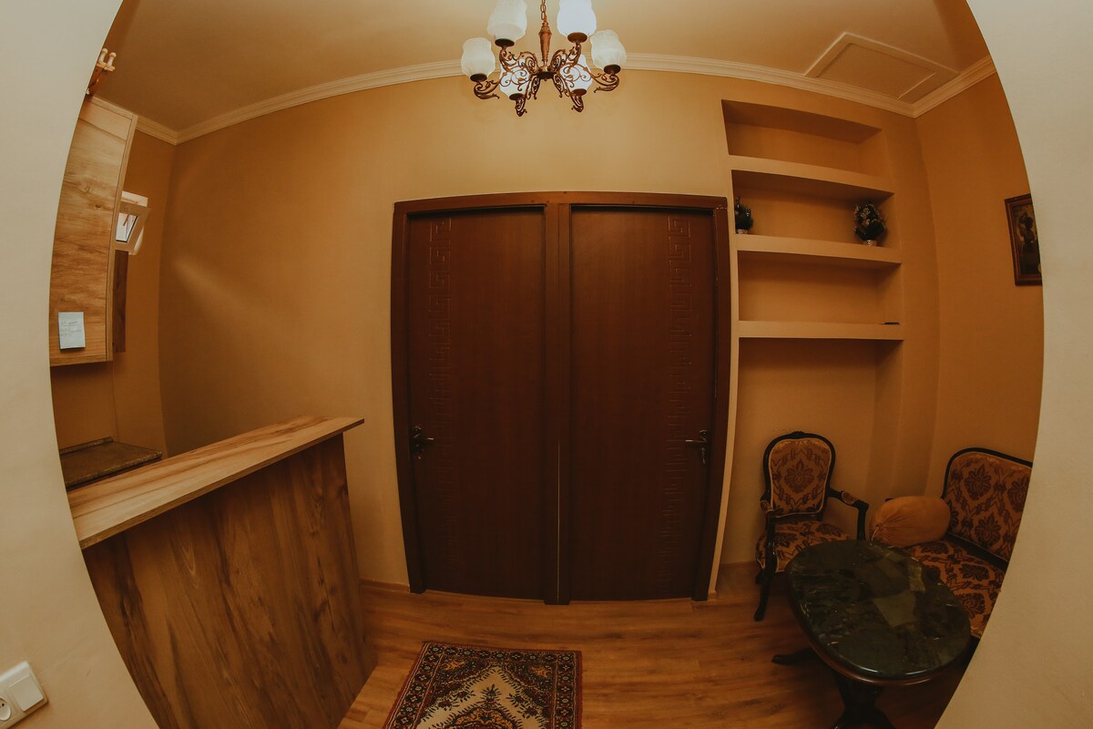 A spacious entrance area is shown, featuring two wooden doors leading to private rooms. A chandelier illuminates the room, while a small seating area with two ornate chairs and a round table is visible. A shelf is mounted on the wall for added storage.
