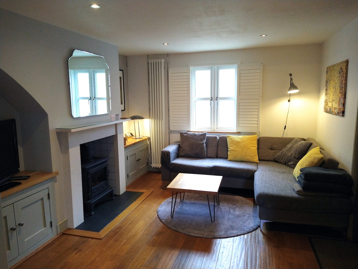 The living area is designed with a corner sofa, accented by cushions. A flat-screen TV is positioned against the wall and a small coffee table sits on a round rug. Light enters through double windows with shutters, and a gas stove is showcased beneath the mantel.