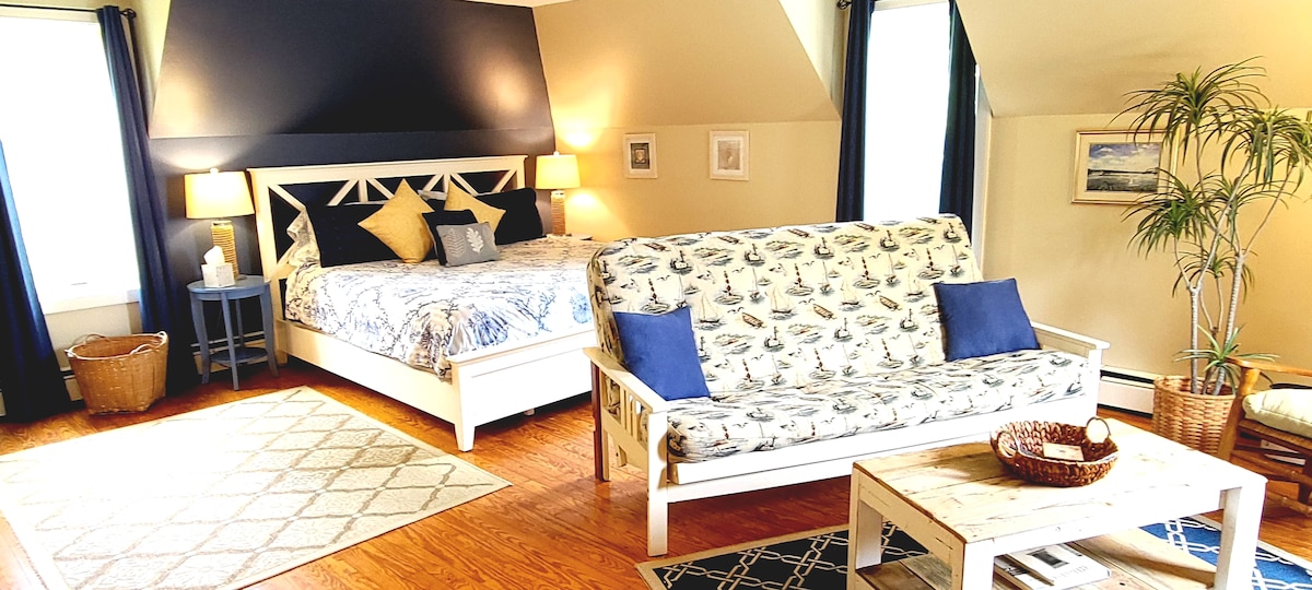 A spacious room features a large king bed with coastal-patterned bedding and a comfortable couch adorned with matching cushions. Natural light floods the space, highlighting the hardwood floors and decorative accents like a plant and framed artwork.