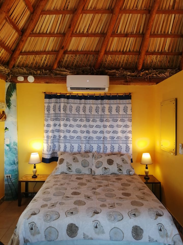 Una cama tamaño queen muy cómoda debajo del aire acondicionado mini split de dos toneladas. Las ventanas están adornadas con hermosas cortinas hechas a mano.