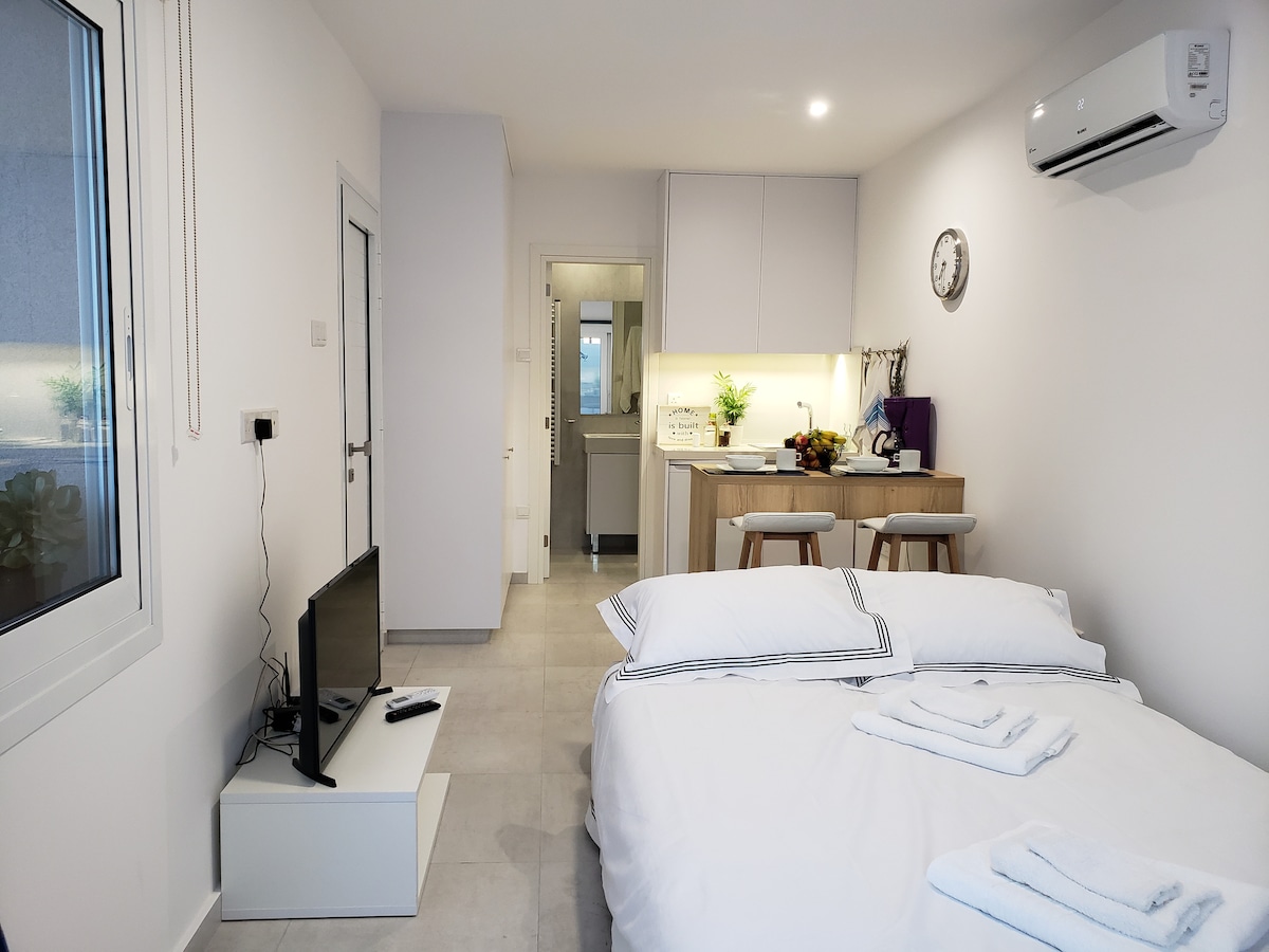 A well-lit studio space features a spacious bed with neatly arranged towels and minimalist decor. A small kitchenette and dining area are visible in the background. The room includes modern amenities, such as a wall-mounted television and air conditioning unit.