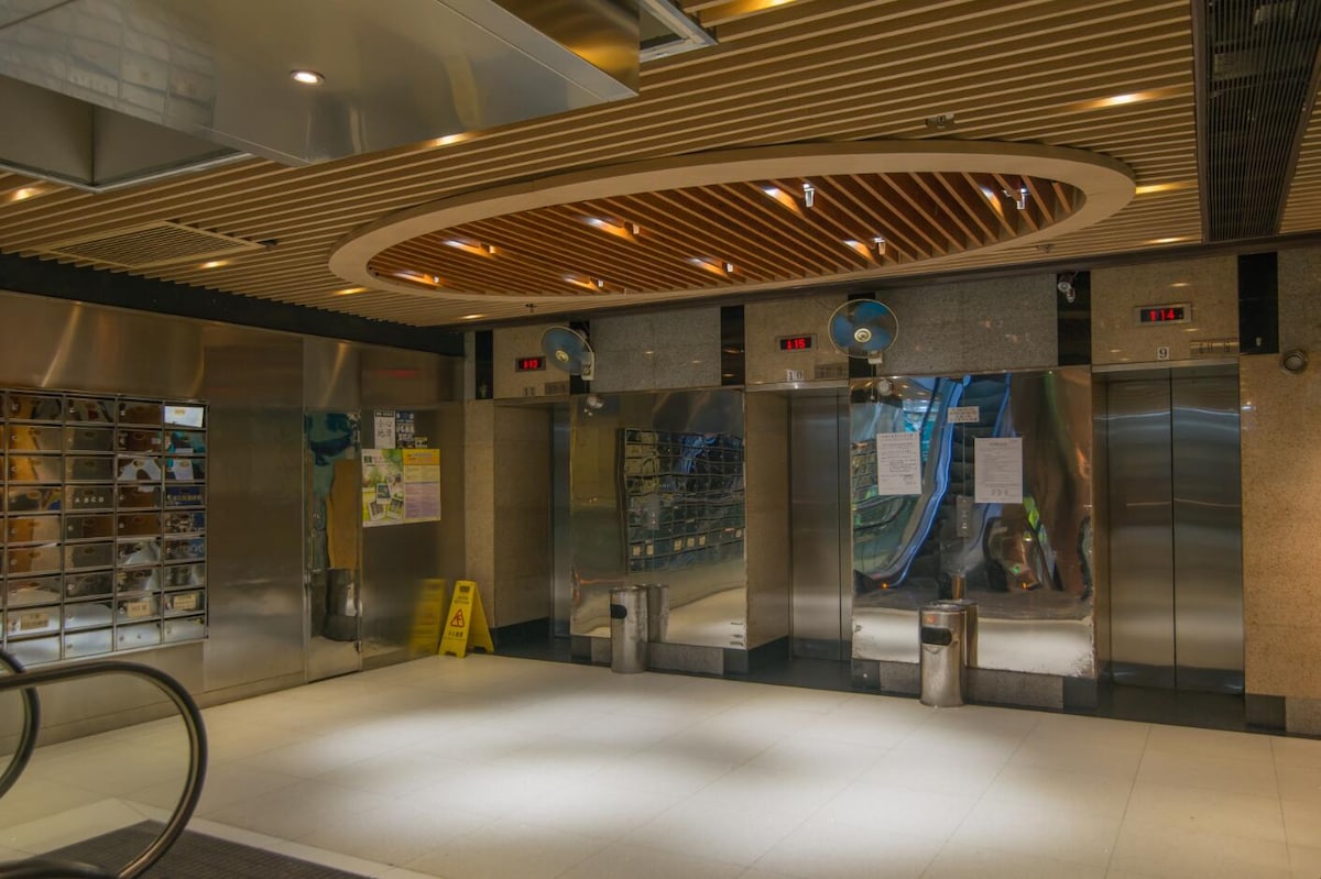 The lobby area features a modern design with polished metallic surfaces and a wooden ceiling. Multiple elevators are visible, accompanied by informational signs. A caution sign is placed on the floor, indicating ongoing maintenance or caution in the area.