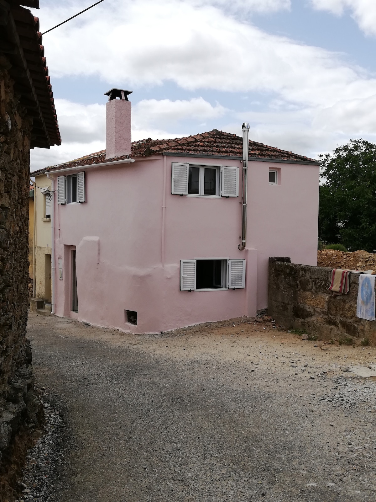 A charming pink house is situated at the end of a gravel road. The property features white shuttered windows and a chimney, with a small opening visible on the lower level. Surrounding landscapes include rustic stone walls and greenery.