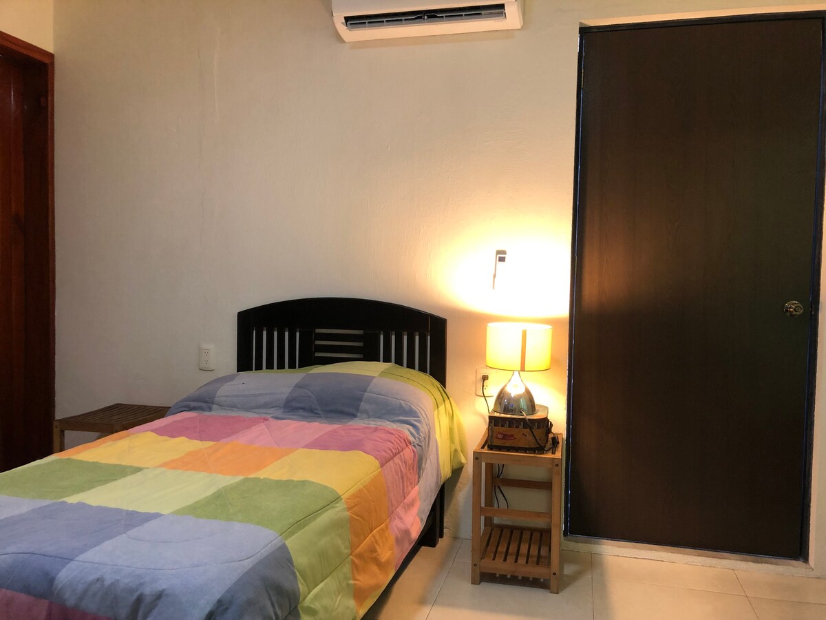 A colorful bedspread covers a single bed, positioned next to a wooden nightstand with a lamp. The room features a simple design, with a wall-mounted light and a closet door that provides privacy.