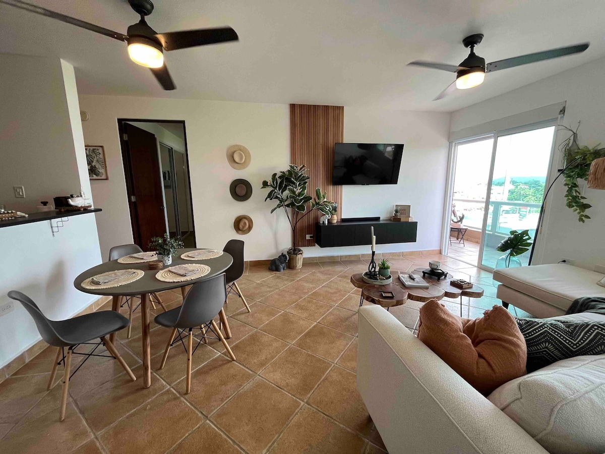 A living area is presented with a round dining table surrounded by six chairs, and a cozy sofa with cushions. A mounted flat-screen TV is visible on the wall, complemented by decorative plants. Natural light enters through sliding glass doors leading to a balcony.
