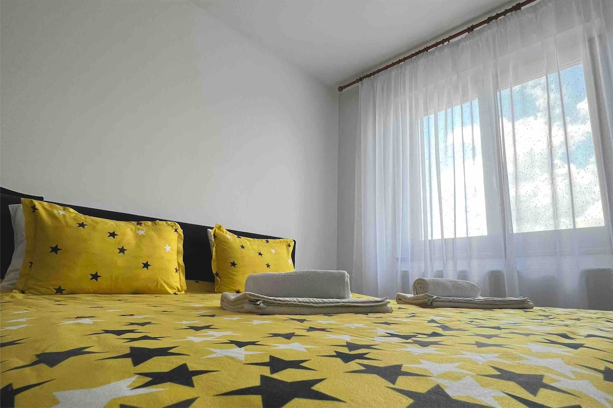 A bright bedroom features a spacious bed adorned with yellow bedding decorated with stars. Two rolled towels are neatly placed on the bed's edge. Natural light filters through the sheer white curtains, illuminating the room.