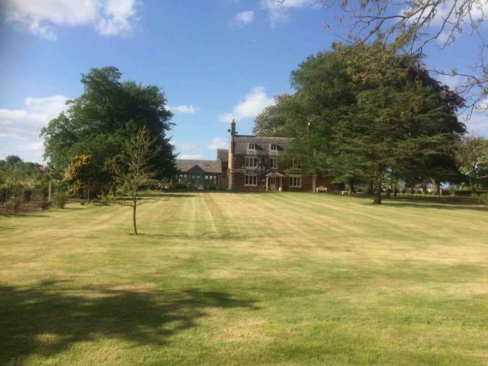 A well-maintained lawn surrounds the Old Coach House, framed by mature trees. The charming brick property features large windows and multiple stories, reflecting the warm daylight. The expansive, open space invites guests to enjoy the serene countryside atmosphere.