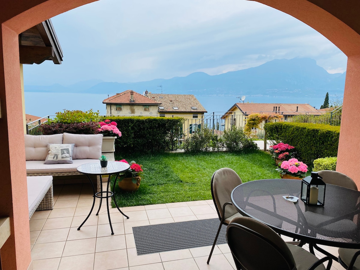 An outdoor terrace is shown, featuring a round table with chairs and a small lounge area with a cushioned sofa. Lush greenery and colorful flowers frame the space, with a panoramic view of Lake Garda and the surrounding mountains visible in the distance.