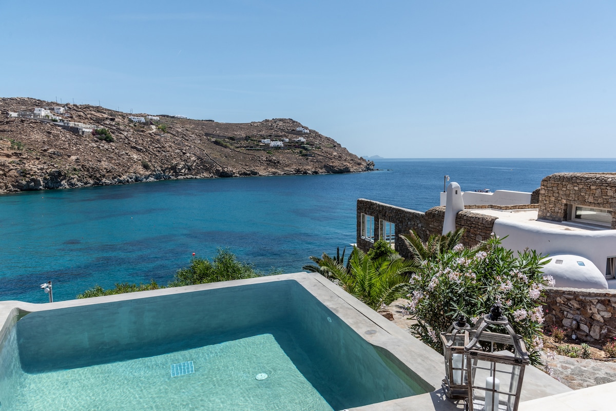A private pool is seen with turquoise water, overlooking the expansive sea and rocky coastline. Lush greenery and villa architecture frame the scene, with a clear blue sky above, enhancing the serene beachfront setting.