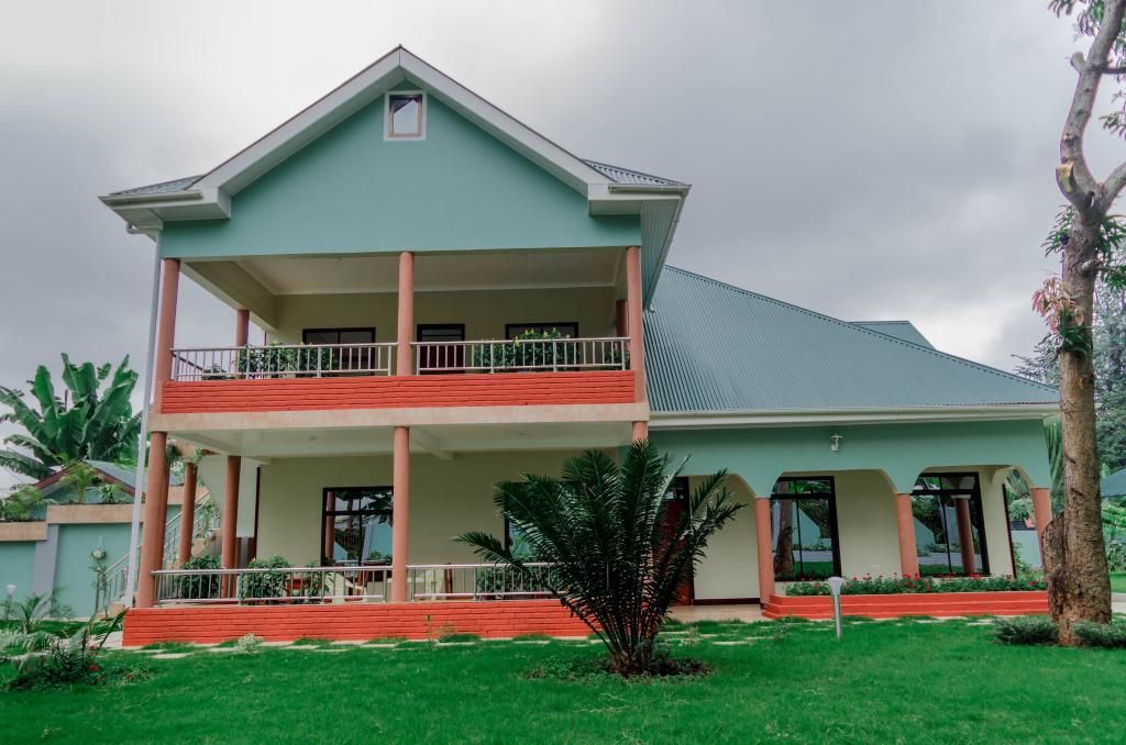 The two-story house displays a combination of light blue and white exterior walls, with distinctive red accents along the base. A spacious porch extends across the front, supported by columns. Lush greenery surrounds the property, complemented by manicured lawns and decorative plants.