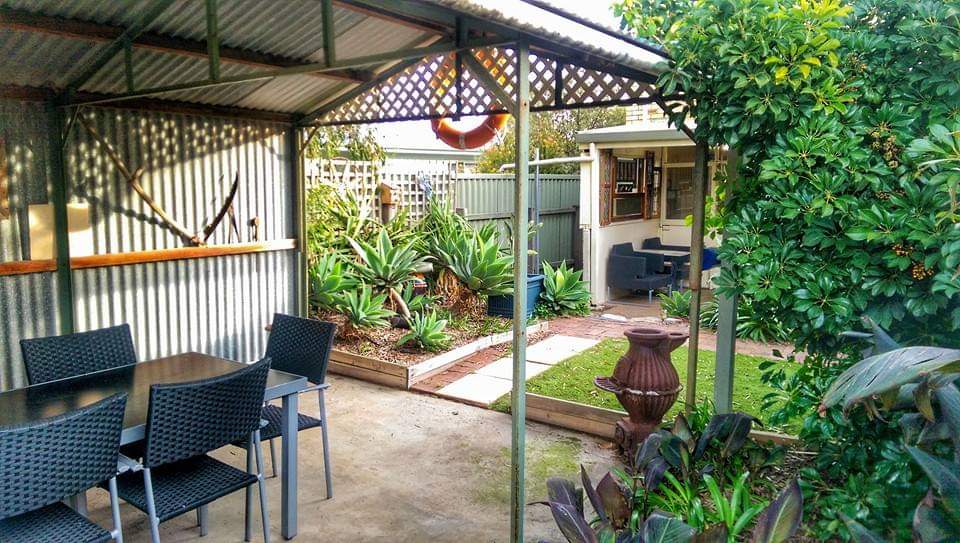 An outdoor dining area features a metal table surrounded by black woven chairs, sheltered under a gazebo. Lush greenery and planters are visible in the background, with a cozy seating area and a decorative pot enhancing the tranquil garden setting.