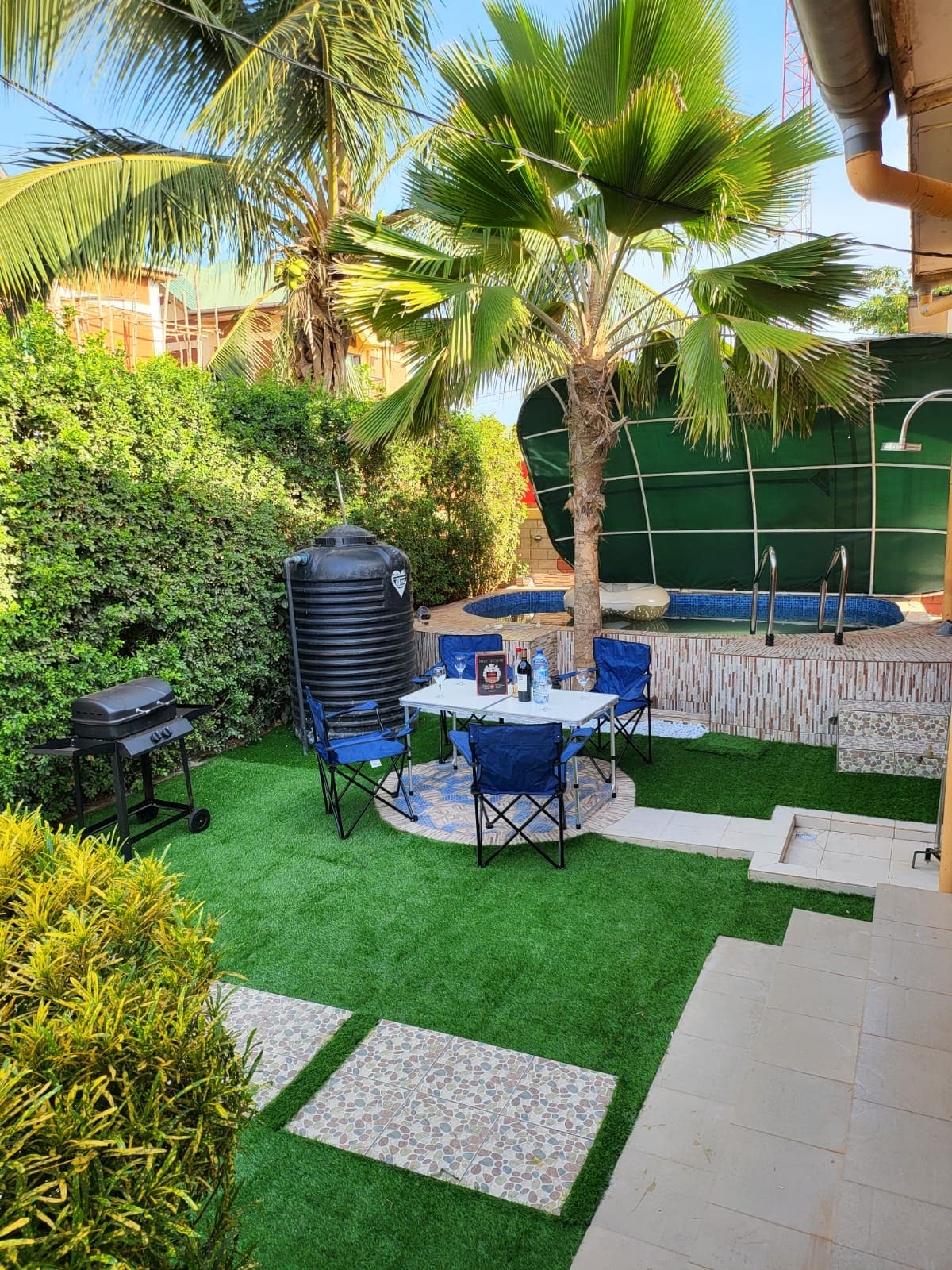An outdoor space is depicted, featuring grass-like flooring, surrounded by lush greenery. A round table with four blue chairs is visible near a barbecue grill. A small swimming pool is located in the background, with a palm tree providing shade.