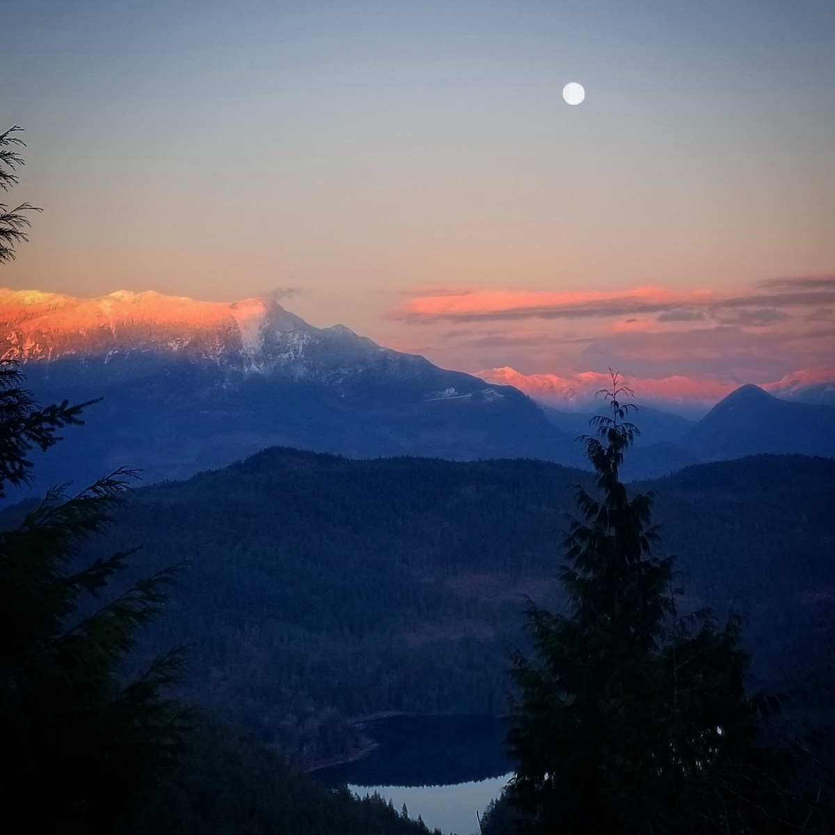 The image features a mountainous landscape bathed in soft hues of orange and pink as the sun sets, with snow-capped peaks visible in the distance. A bright moon is positioned in the sky, illuminating the scene while a body of water reflects the surrounding nature below.