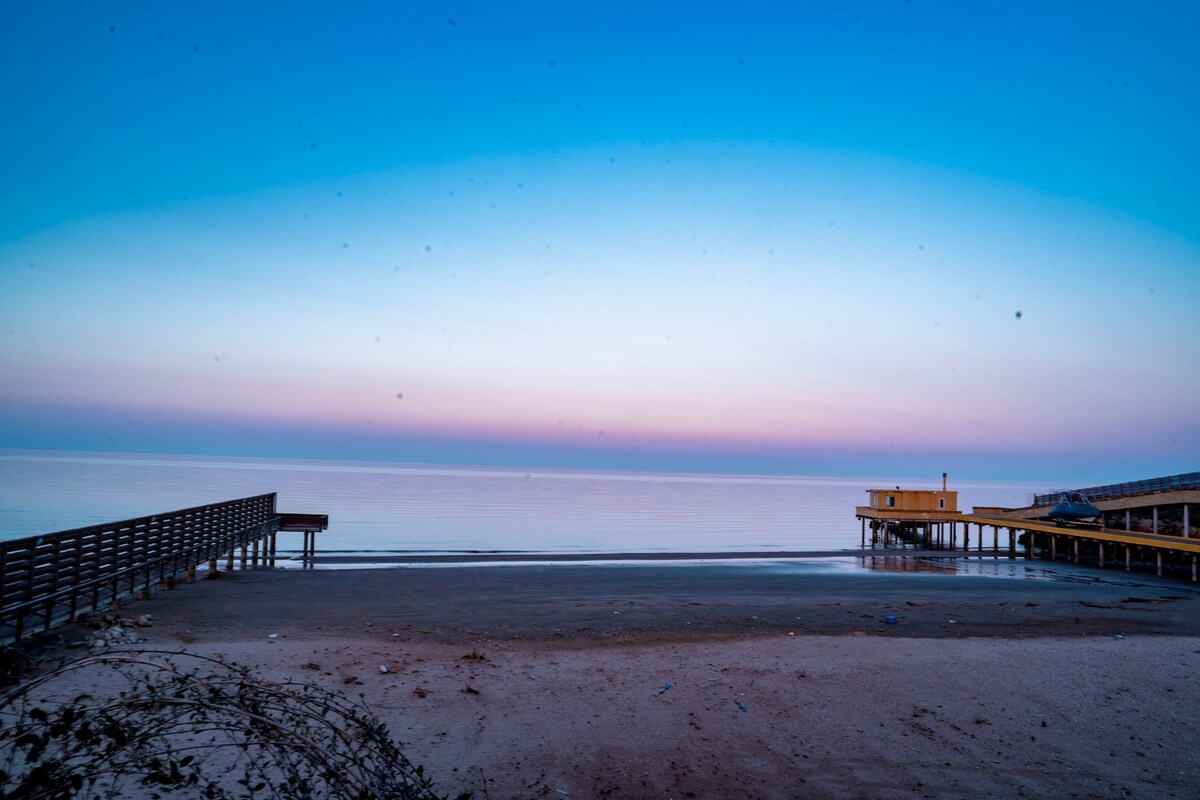 A serene beach scene is depicted, showcasing calm waters meeting a pastel sky at dawn. Wooden piers extend into the sea, and soft sand stretches across the foreground, providing a tranquil setting for relaxation.