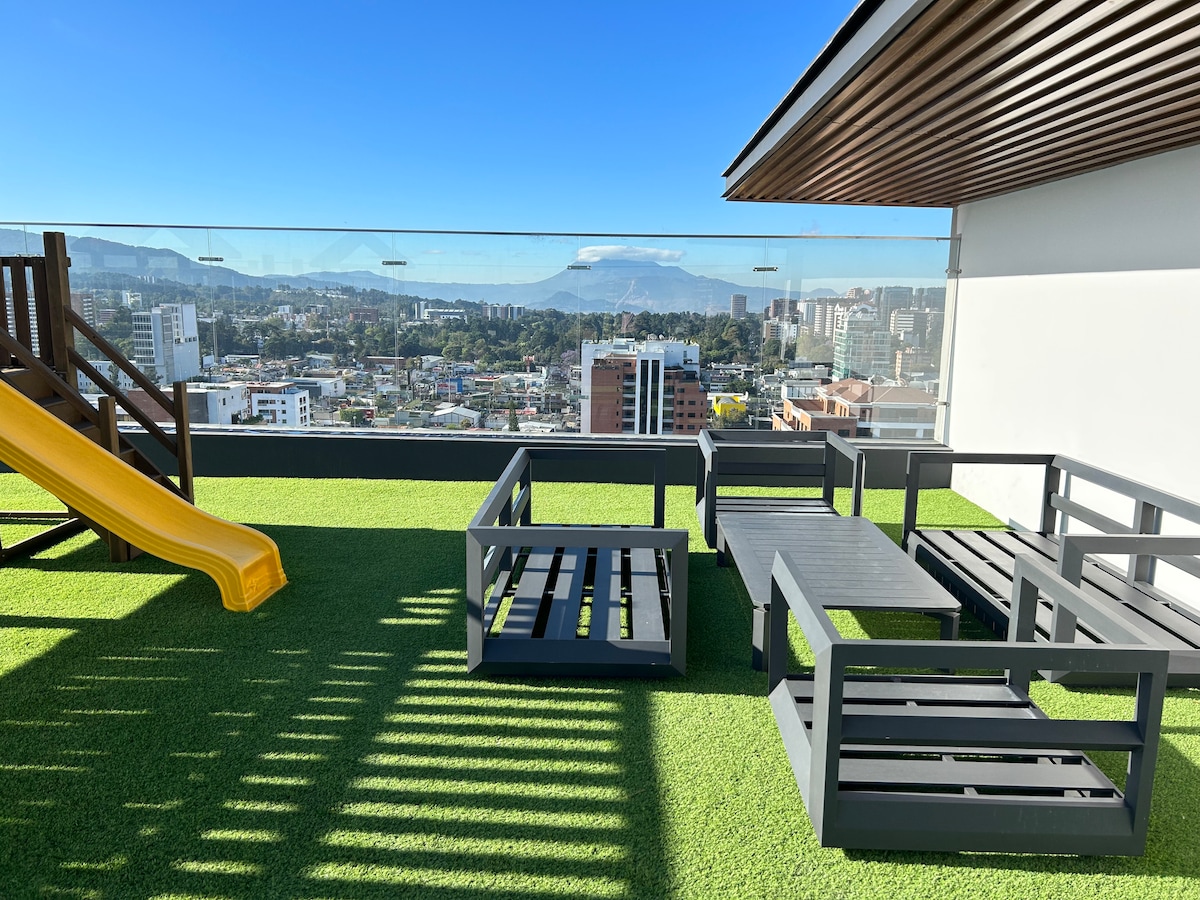 An outdoor rooftop space features synthetic grass, offering comfortable seating arrangements made of gray wooden frames. A yellow slide extends from the left side, providing a fun option for children. The area is surrounded by glass panels, revealing expansive views of the city and surrounding mountains.