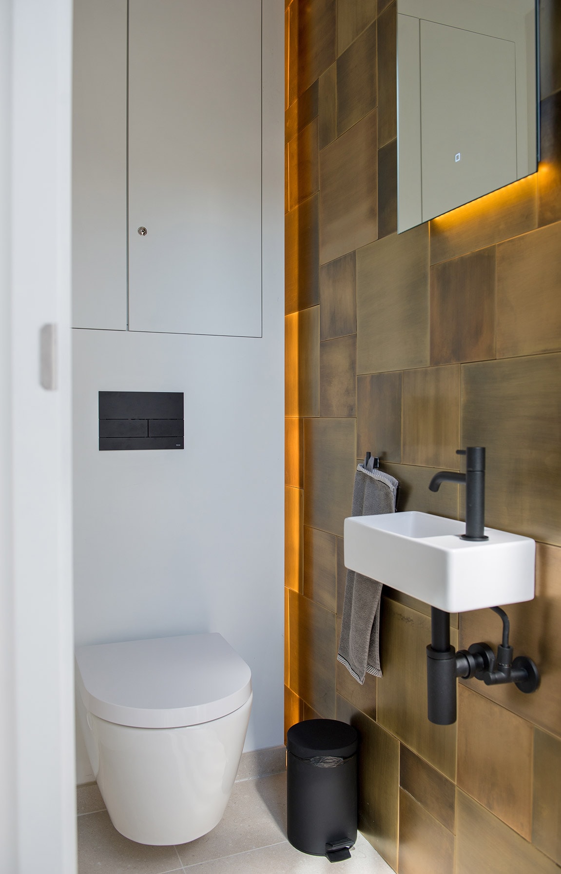A modern restroom is presented, featuring a wall-mounted toilet and a sleek sink with a matte black faucet. The wall is adorned with textured bronze tiles, illuminated by soft lighting. A small mirror hangs above the sink, and a towel is neatly hung by a hook.