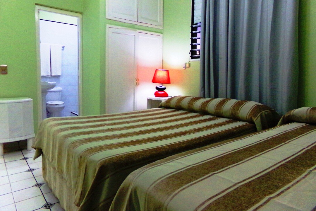 A room featuring two beds with striped bedding, placed beside a bedside table with a small lamp. Neutral green walls and light curtains create a soft atmosphere. A bathroom is visible through an open door, and ample storage is provided by a wardrobe.