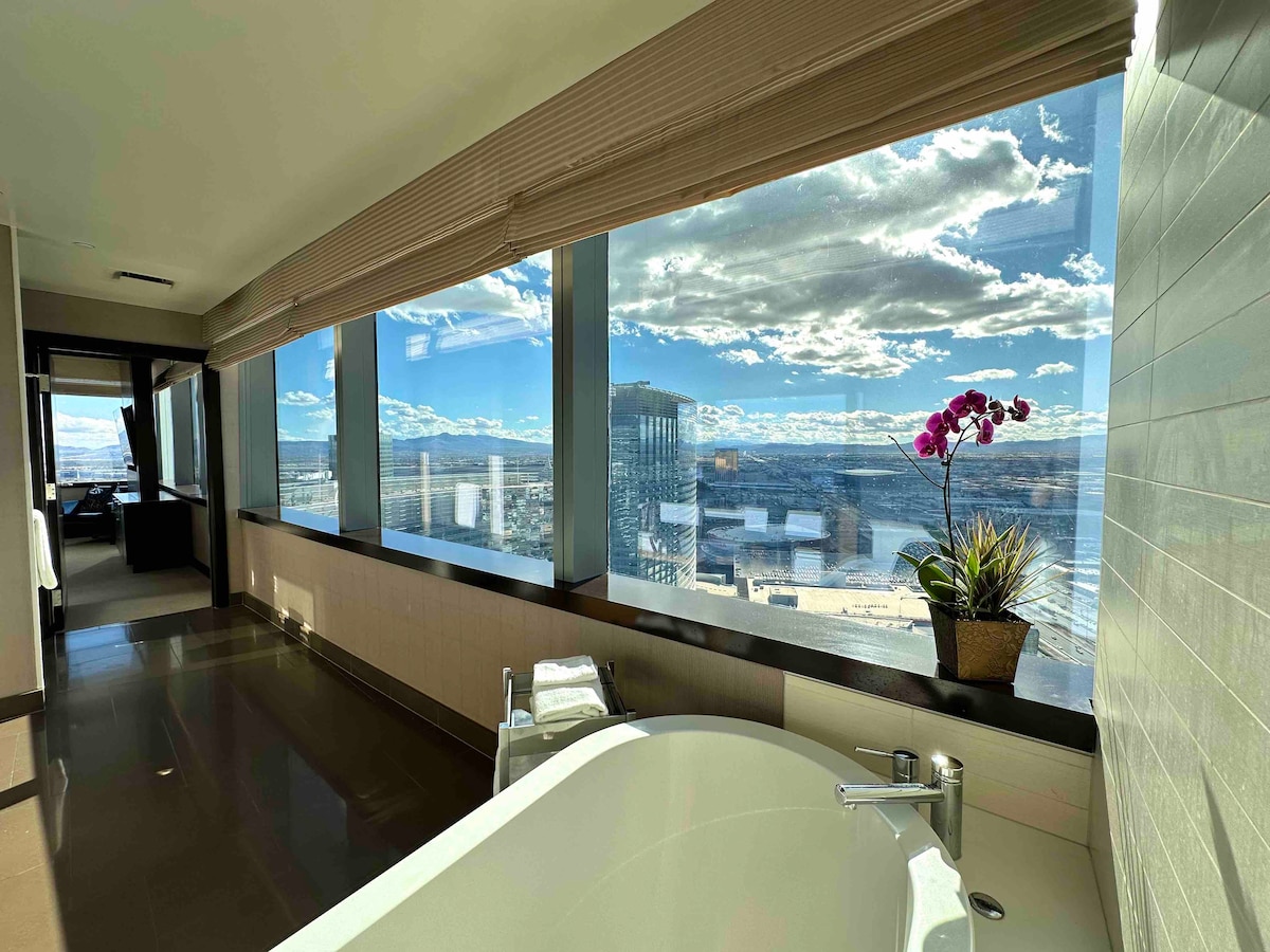 A spa-style bathroom features a deep soaking tub positioned near large windows that offer panoramic views of the surrounding landscape. Natural light illuminates the space, enhanced by a potted orchid and towels neatly arranged on the counter.