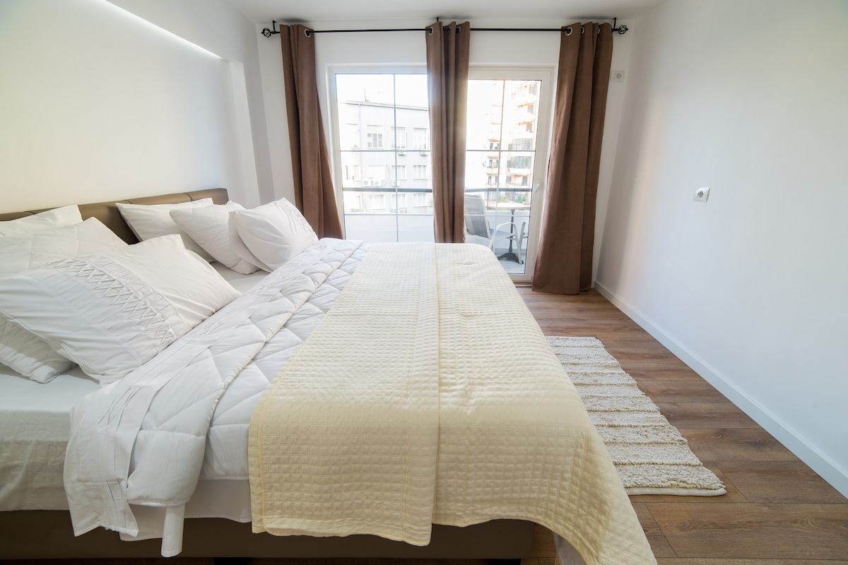 A spacious bedroom is illuminated by natural light from a large window, featuring a comfortable bed adorned with neatly arranged pillows and a soft blanket. A light-colored area rug complements the parquet flooring, while elegant curtains frame the window, enhancing the serene environment.