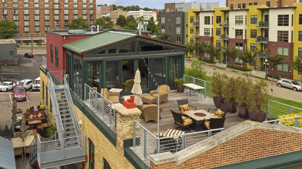A rooftop patio features comfortable seating arrangements and a central fire pit, offering a social gathering space with views of the surrounding urban landscape. Potted plants enhance the area, while a glass-enclosed sunroom provides indoor access and an open design that invites natural light.