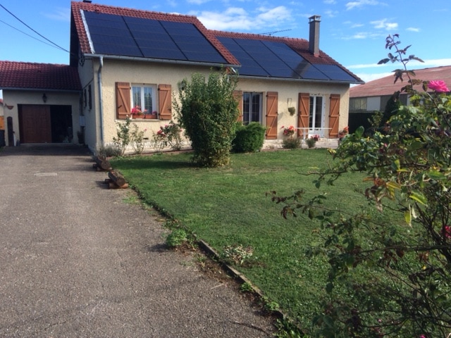 A charming country house is seen, featuring a light exterior with wooden shutters. Solar panels are mounted on the roof. The landscaped garden includes neatly trimmed grass and blooming flowers, providing a peaceful outdoor environment. A gravel pathway leads to the home.