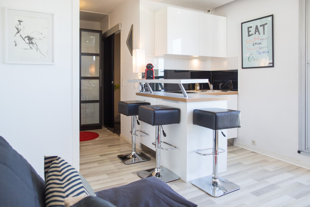 A modern kitchen area is equipped with sleek, white cabinetry and black countertops. Three bar stools with black upholstery are positioned at a white island, providing a casual dining spot. Light wood flooring complements the contemporary design and enhances the open feel of the space.
