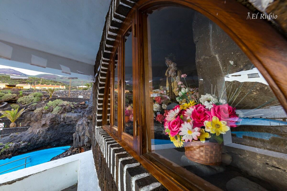 A stone archway frames a window showcasing a basket of colorful artificial flowers in vibrant hues. The surrounding landscape features volcanic rock and a glimpse of blue water, suggesting a serene outdoor environment nearby.