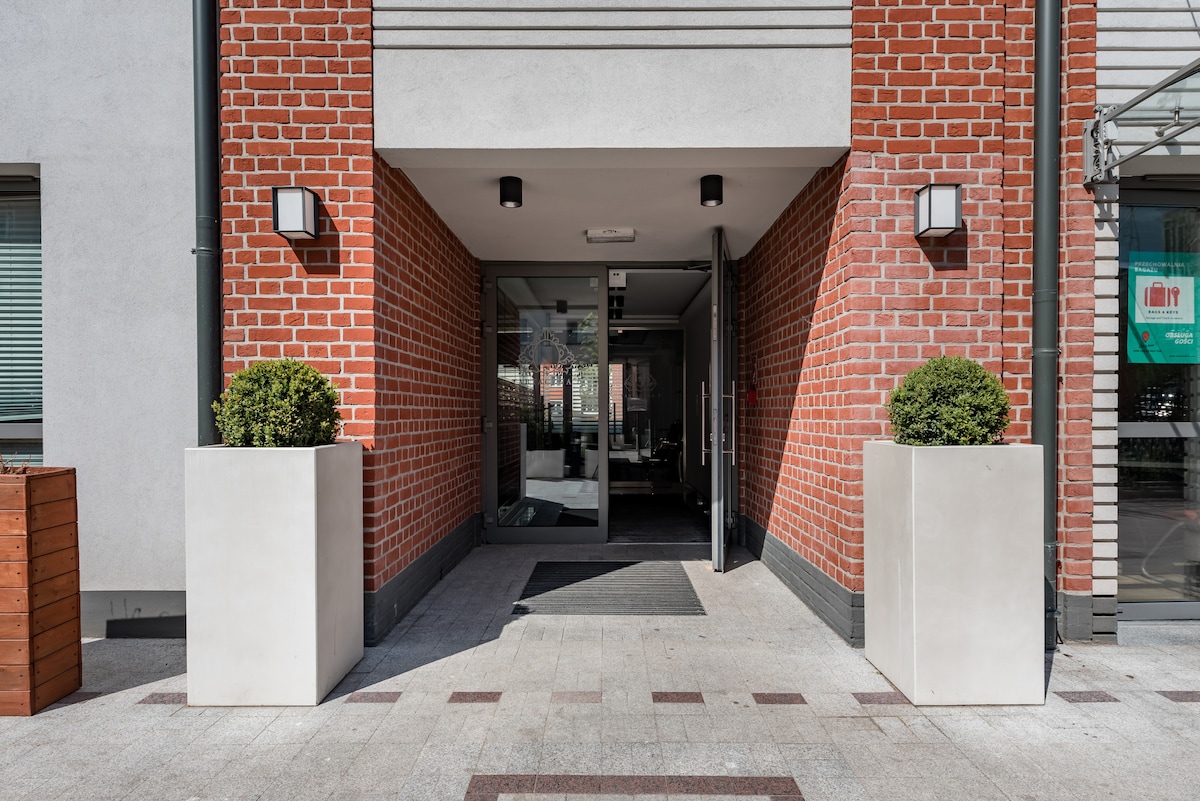 The entrance features a modern design with a smooth pathway flanked by tall, minimalistic planters. Brick walls create a warm facade, while wall sconces provide subtle illumination at the entrance, leading to clear glass doors that open into the building.