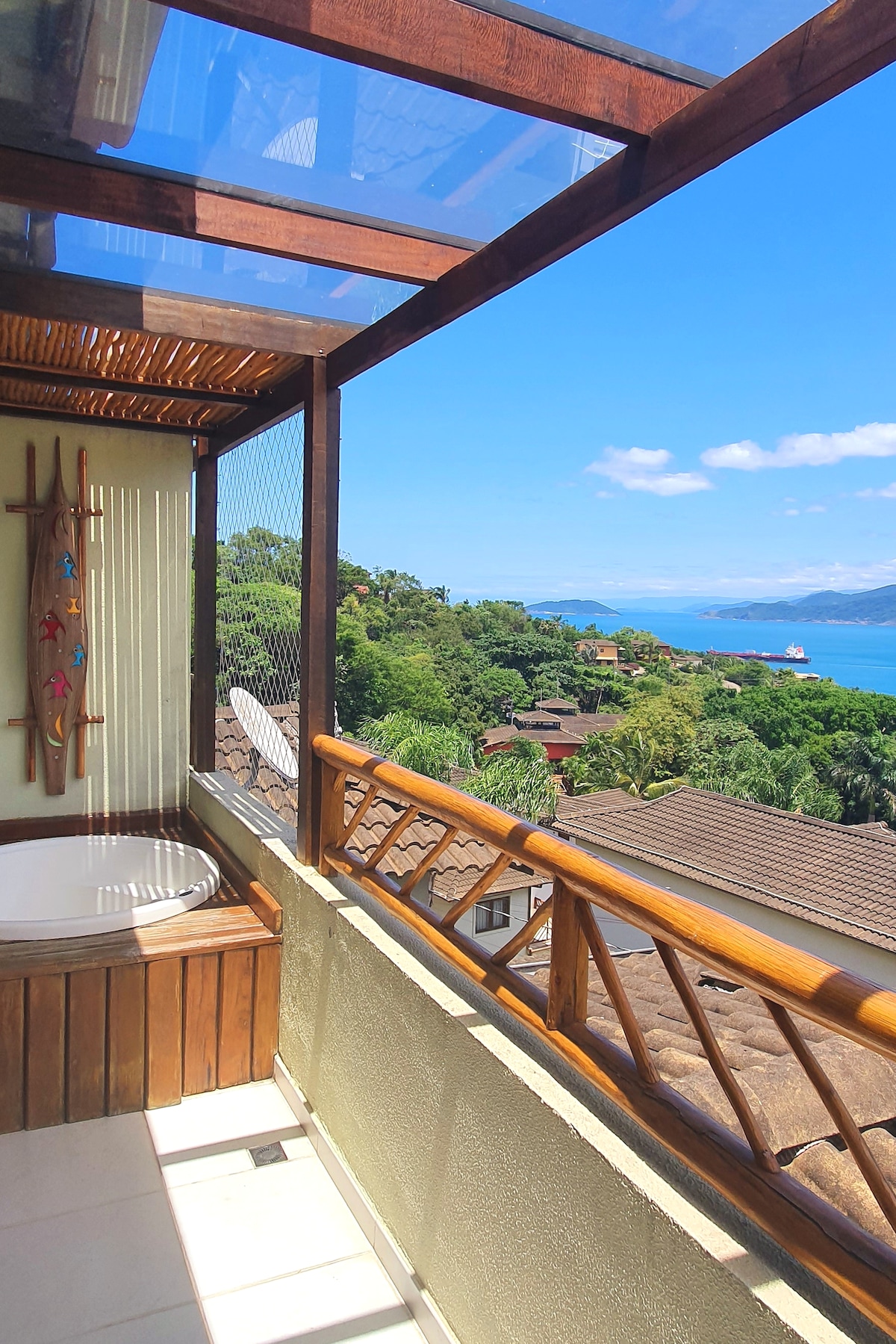 A private balcony features a jacuzzi, providing a view of the lush hillside and sea. The area is partially enclosed with wooden railings, allowing for scenic observation. Natural light illuminates the space, enhancing the inviting atmosphere of the outdoor setting.
