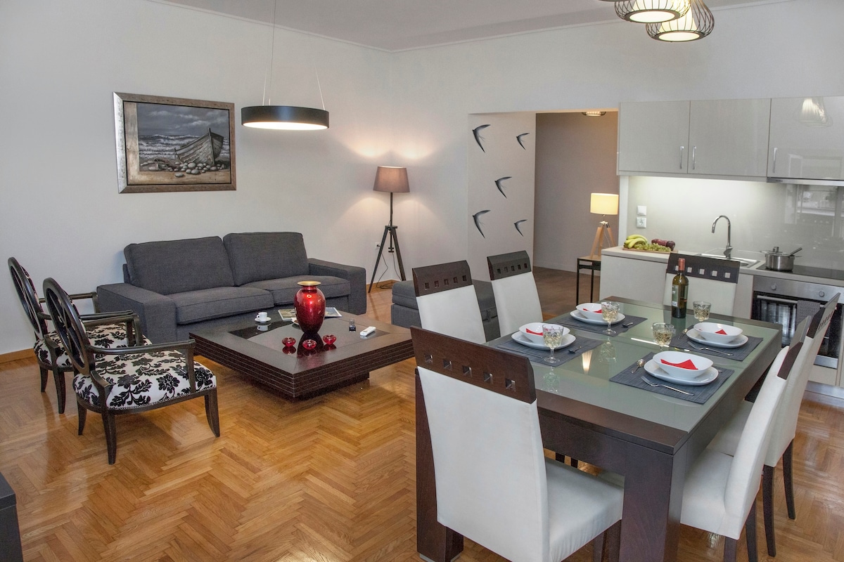 An open living and dining area features a modern dining table set for six, complemented by a stylish coffee table and a plush sofa. A well-equipped kitchen is visible in the background, while soft lighting fixtures enhance the bright space.
