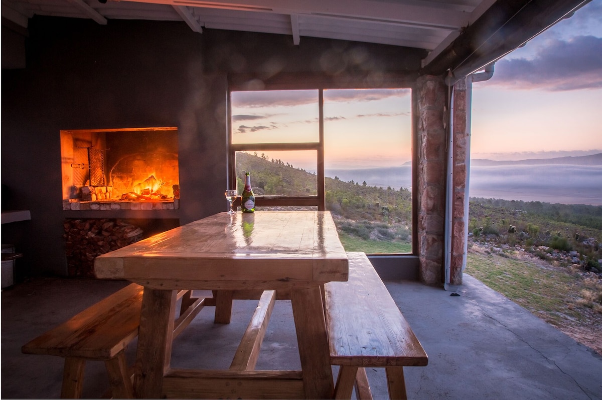 An indoor dining area features a large wooden table with benches, positioned near a stone fireplace. Large windows offer expansive views of the landscape. A bottle of wine and two glasses are placed on the table, with soft light streaming in from the sunset beyond.