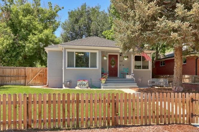The exterior of a modern cottage features a welcoming porch with colorful chairs and a decorative wreath on the front door. A fenced yard with manicured grass and a backdrop of trees adds to the inviting appearance. A pathway leads up to the home.
