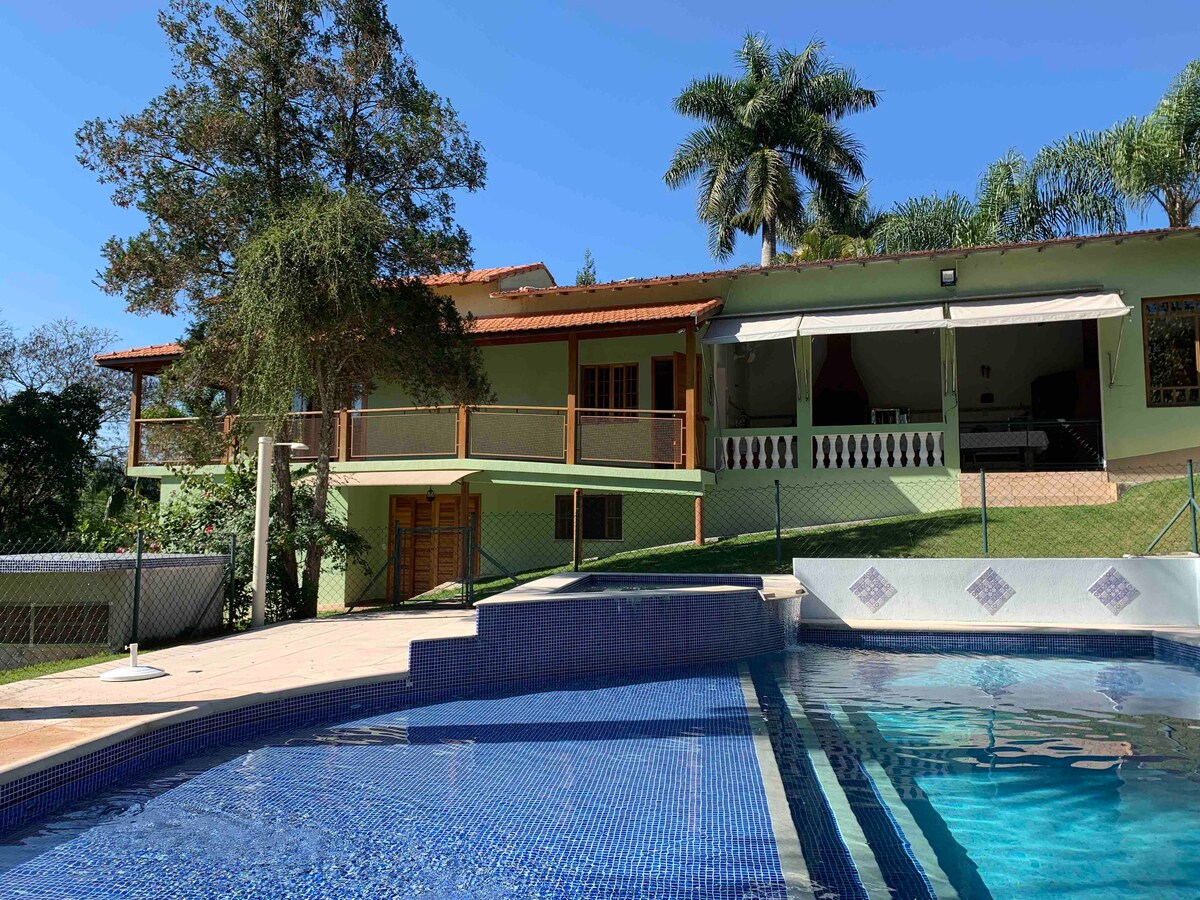 A spacious house is visible above a blue-tiled swimming pool, surrounded by greenery. The exterior includes large balconies and a covered patio area. Palm trees and well-maintained gardens can be seen on the property, enhancing the tranquil outdoor environment.