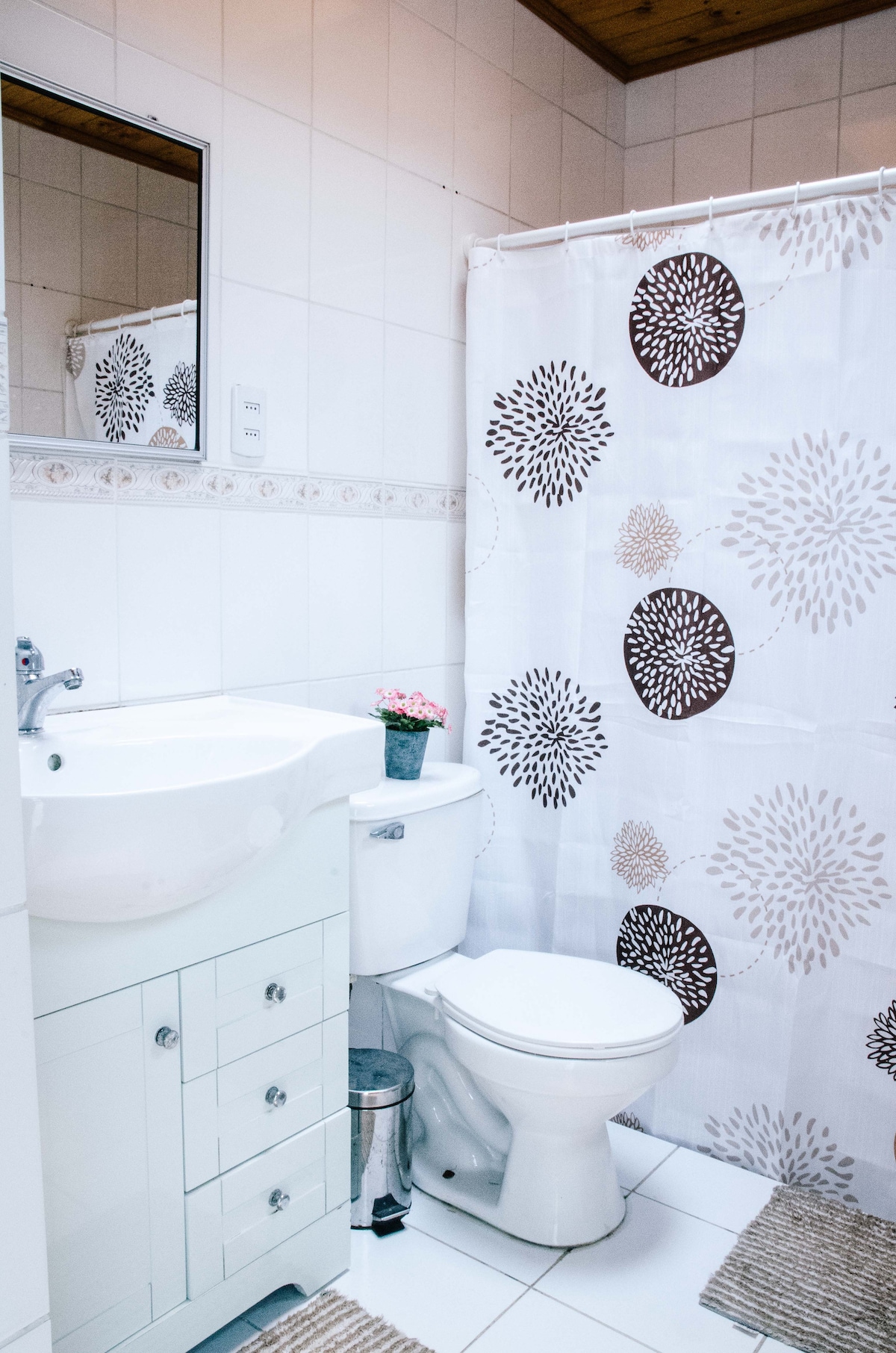 A clean bathroom is depicted, featuring a white sink with a cabinet below. A white toilet is placed beside it, while a patterned shower curtain adds visual interest. A small potted plant sits on the sink, and decorative rugs are laid on the tiled floor.