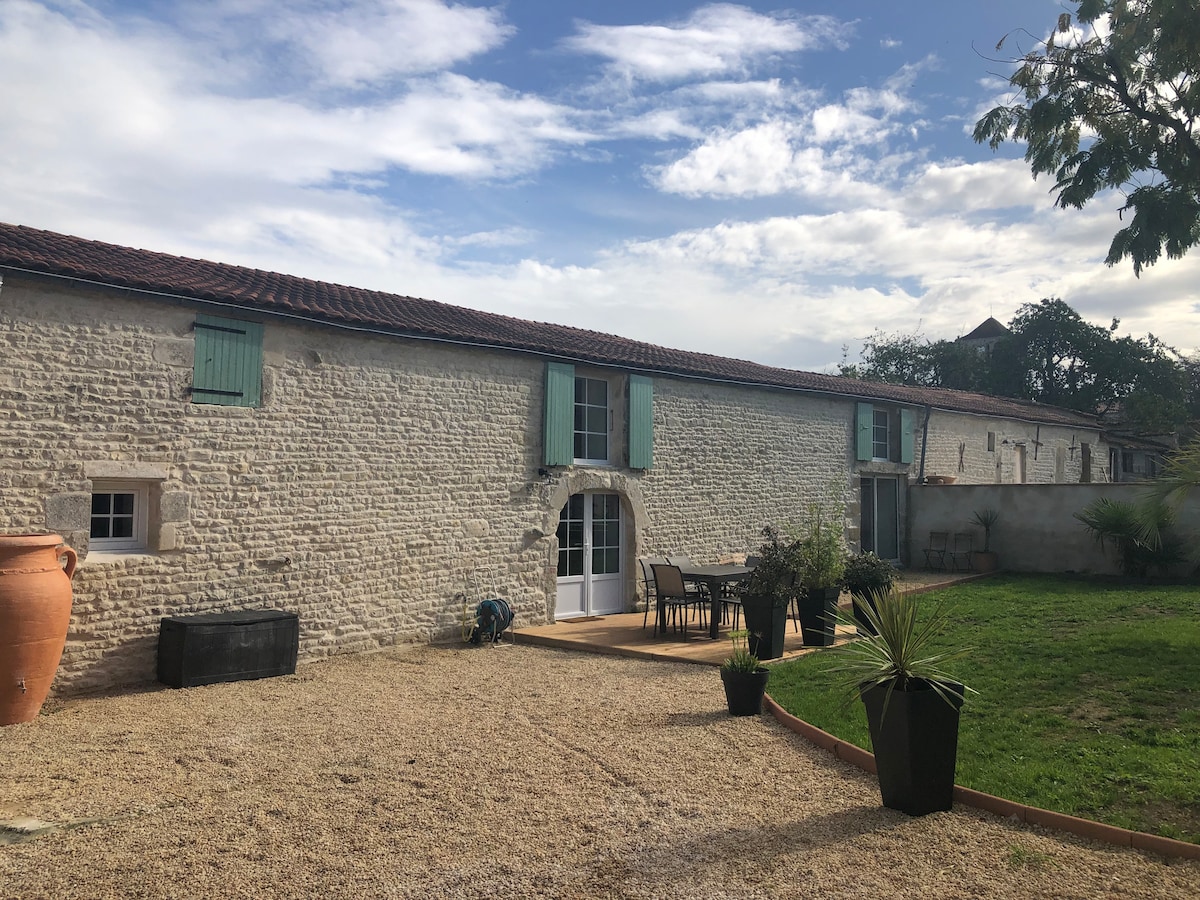 A renovated barn is visible, characterized by light-colored stone walls and teal shutters. The outdoor area features a spacious wooden deck with a dining table and chairs. Lush greenery and potted plants surround the space, providing a pleasant view under a partly cloudy sky.