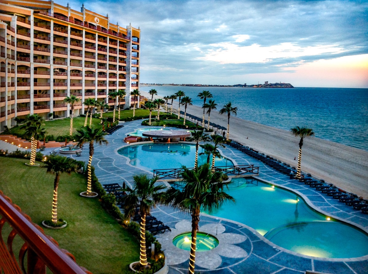 A stunning view of the resort's pool area is depicted, surrounded by lush palm trees. Lounge chairs are arranged poolside, with the beach and ocean visible in the background. The tranquil waters reflect the sky, showcasing the peaceful atmosphere of the beachfront retreat.