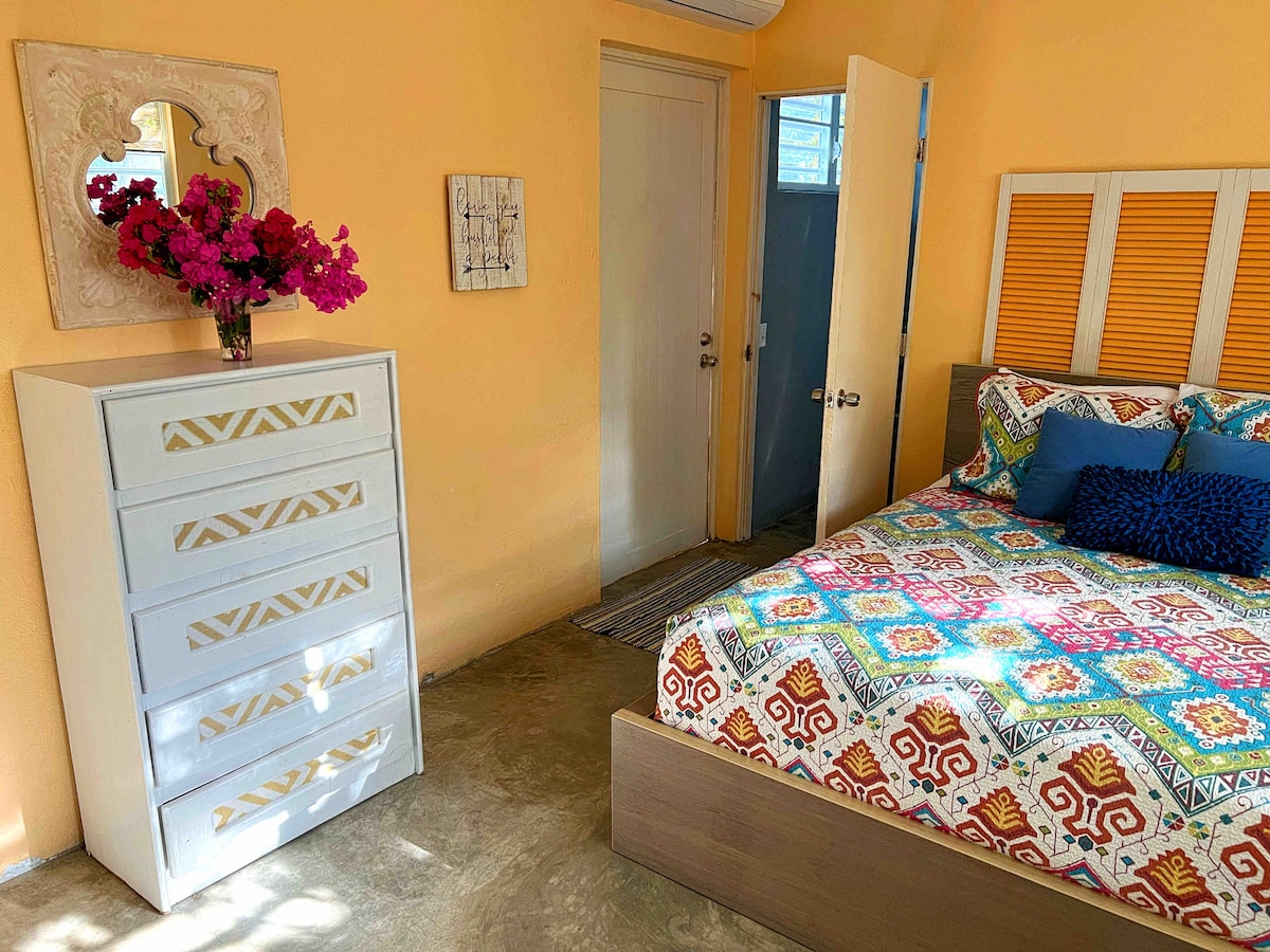 A bright bedroom features a queen size bed adorned with a colorful patterned bedspread and accent pillows. A chest of drawers stands nearby, topped with a vase of vibrant pink flowers. An open door leads to a private bathroom, illuminated by natural light.