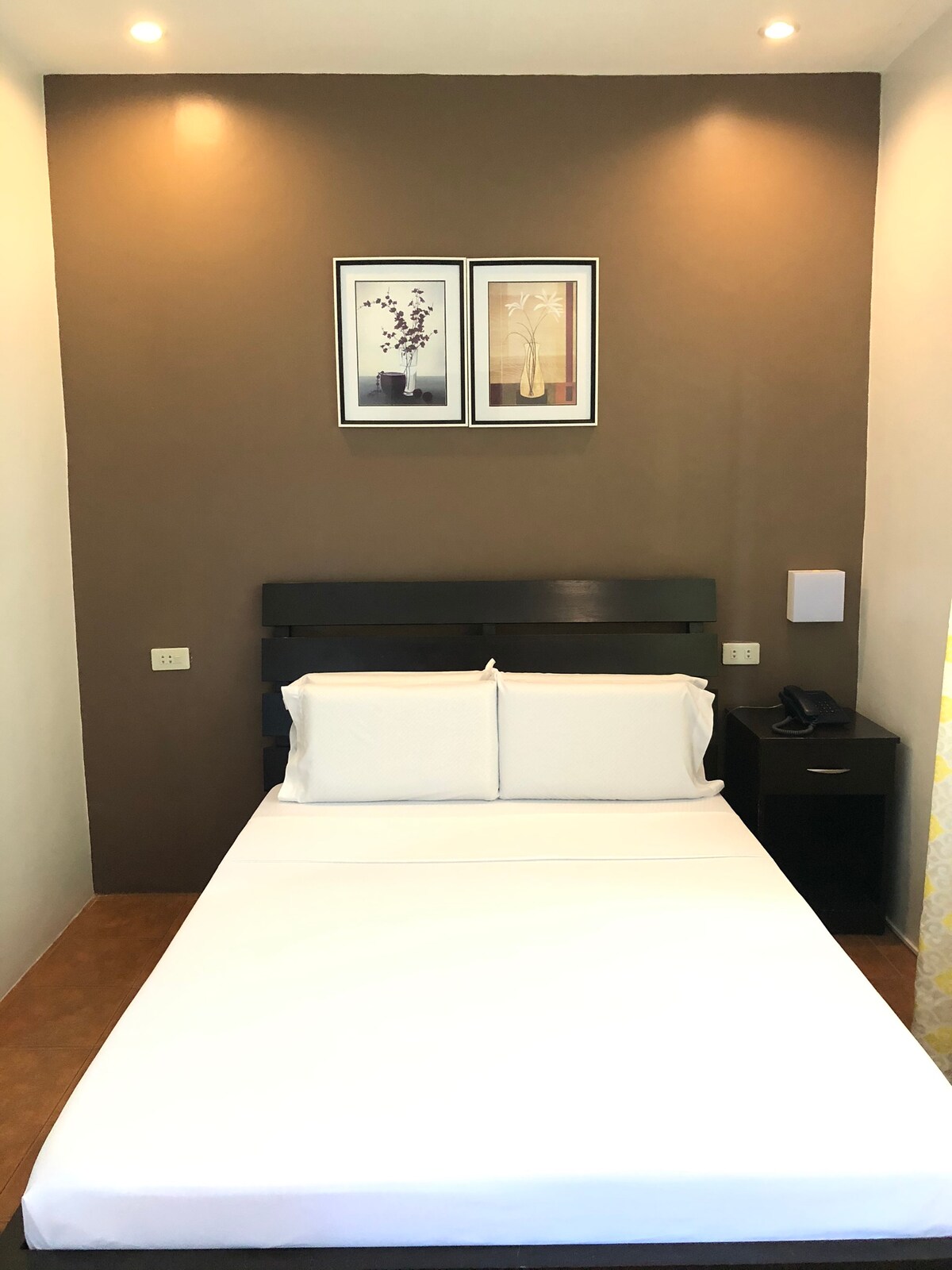 A neatly arranged sleeping area features a spacious bed with white linens against a dark accent wall. Two framed artworks adorn the wall above the bed. A bedside table is positioned alongside, with a phone and a lamp prominently placed.